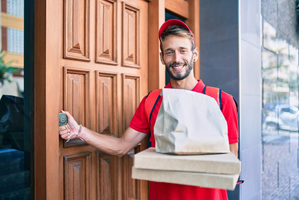 Kaukaski dostawca w czerwonym mundurze i plecaku dostawy uśmiechnięty szczęśliwy na zewnątrz trzymając pudełko pizzy - Zdjęcie, obraz