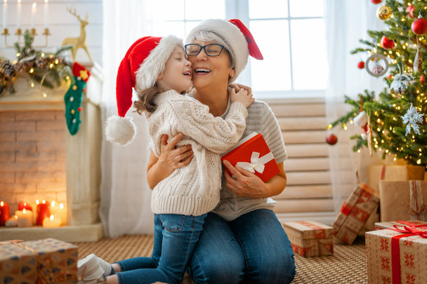 Merry Christmas and Happy Holidays! Cheerful grandma and her cute grand daughter girl exchanging gifts. Granny and little child having fun near tree indoors. Loving family with presents in room. - Foto, Imagem