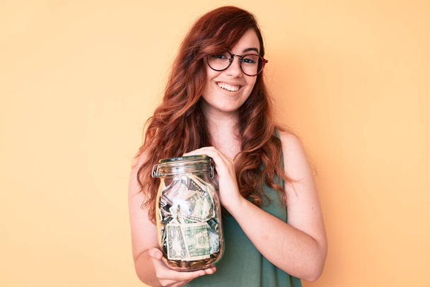 Young beautiful woman wearing glasses holding jar with savings looking positive and happy standing and smiling with a confident smile showing teeth  - Фото, зображення