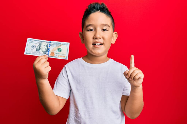 Маленький мальчик латиноамериканец с банкнотой в 100 долларов, улыбающийся идеей или вопросом, указывающий пальцем со счастливым лицом, номер один  - Фото, изображение