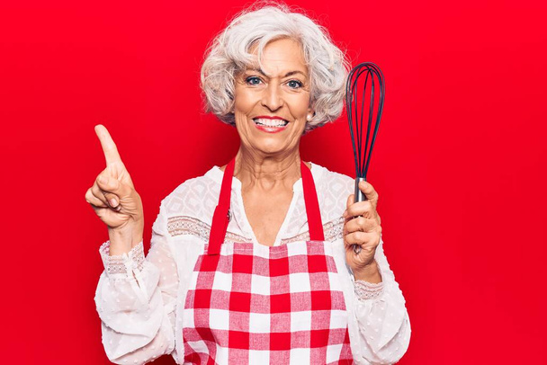 Senior grey-haired woman wearing apron holding whisk smiling happy pointing with hand and finger to the side  - Valokuva, kuva