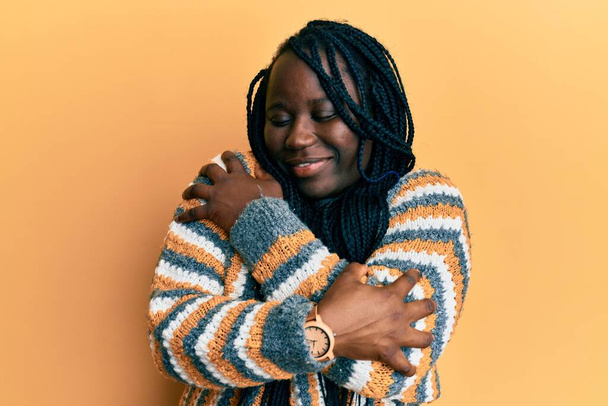 Jonge zwarte vrouw met vlechten dragen casual winter trui knuffelen zichzelf gelukkig en positief, glimlachend zelfverzekerd. zelfliefde en zelfzorg  - Foto, afbeelding