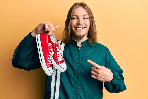Handsome caucasian man with long hair holding red casual shoes smiling happy pointing with hand and finger  - Photo, Image