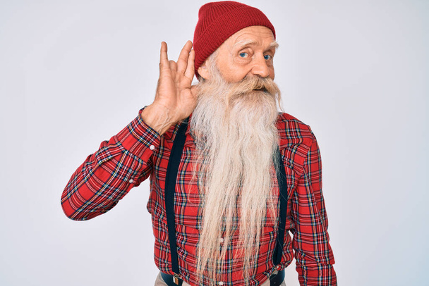 Γέρος ηλικιωμένος άνδρας με γκρίζα μαλλιά και μακριά γενειάδα φορώντας hipster εμφάνιση με μάλλινο καπέλο χαμογελώντας με το χέρι πάνω από το αυτί ακούγοντας μια ακρόαση για φήμες ή κουτσομπολιά. έννοια της κώφωσης.  - Φωτογραφία, εικόνα