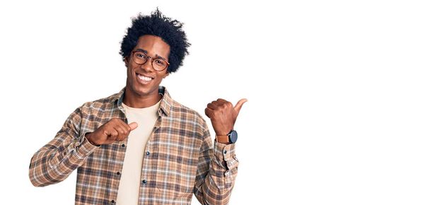 Hezký africký Američan s afro vlasy na sobě ležérní oblečení a brýle ukazuje na zadní straně s rukou a palci nahoru, s úsměvem sebevědomě  - Fotografie, Obrázek