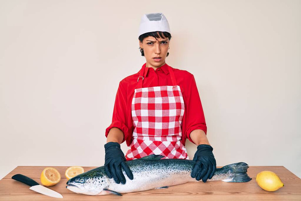 Krásná brunetka žena rybář vaření čerstvé syrové lososa bezradný a zmatený výraz. pochybný koncept.  - Fotografie, Obrázek