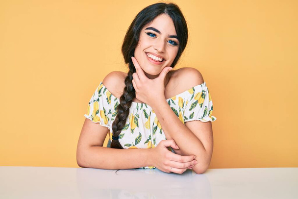 Brunette teenager girl wearing casual clothes sitting on the table looking confident at the camera smiling with crossed arms and hand raised on chin. thinking positive.  - Foto, Imagem
