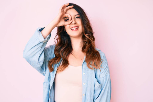 Young beautiful brunette woman wearing casual clothes over pink background smiling happy doing ok sign with hand on eye looking through fingers  - Foto, afbeelding
