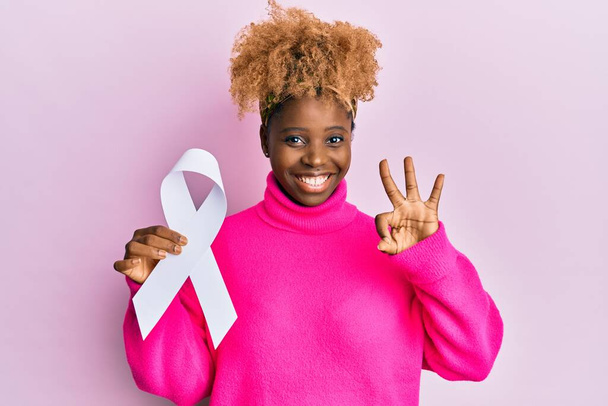 Young african woman with afro hair holding pink cancer ribbon doing ok sign with fingers, smiling friendly gesturing excellent symbol  - Fotografie, Obrázek