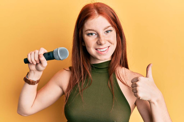 Молода дівчина червоної голови співає пісню, використовуючи мікрофон усміхнений щасливий і позитивний, великий палець робить відмінний знак схвалення
  - Фото, зображення