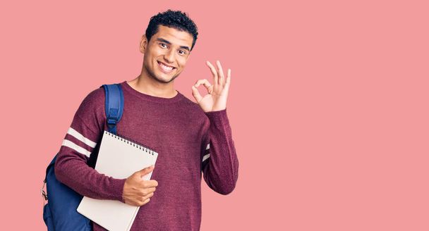 İspanyol yakışıklı genç adam öğrenci çantası ve not defteri takıyor tamam imzasını atıyor parmaklarıyla, gülümseyerek mükemmel bir sembol oluşturuyor  - Fotoğraf, Görsel