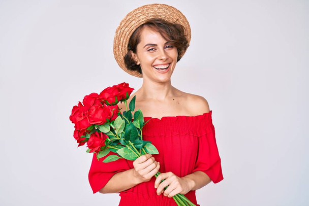 Young hispanic woman wearing hat holding flowers looking positive and happy standing and smiling with a confident smile showing teeth  - Photo, Image