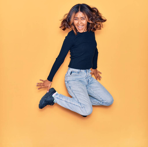 Young beautiful latin woman smiling happy. Jumping with smile on face and arms opened over isolated yellow background - Photo, Image