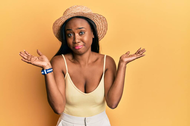 Fiatal afrikai nő visel nyári kalap felett sárga háttér tudatlan és zavaros tárt karokkal, fogalmam sincs, és kétes arc.  - Fotó, kép