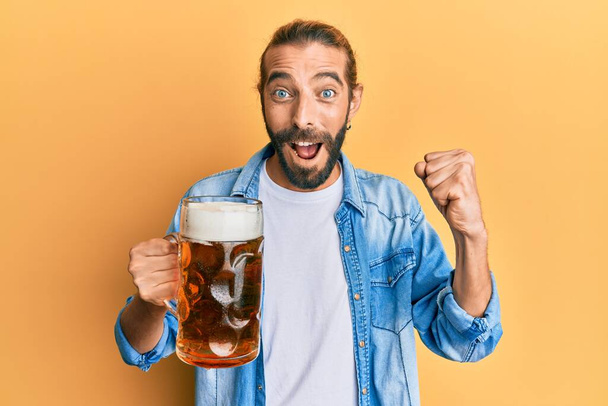 Attraktiver Mann mit langen Haaren und Bart, der stolz ein Pint Bier trinkt und Sieg und Erfolg mit erhobenen Armen feiert  - Foto, Bild