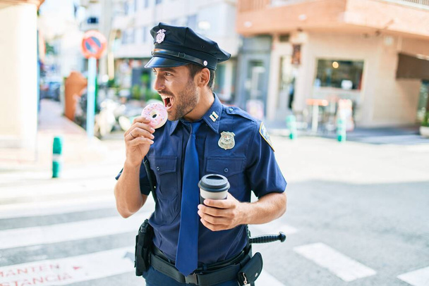 Joven policía hispano guapo vistiendo uniforme de policía sonriendo feliz. Comer rosquilla y beber una taza de café para llevar en la calle de la ciudad. - Foto, Imagen