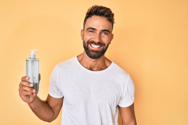 Homem hispânico jovem usando gel higienizador de mão olhando positivo e feliz de pé e sorrindo com um sorriso confiante mostrando dentes  - Foto, Imagem