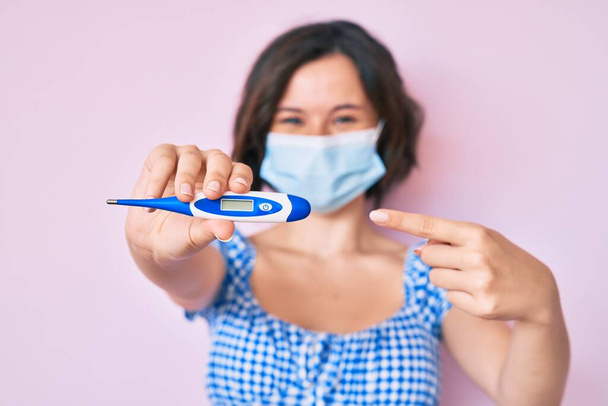 Young beautiful woman wearing medical mask holding holding thermometer smiling happy pointing with hand and finger  - Photo, Image