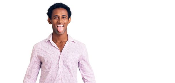 Hombre guapo africano con camisa rosa casual sacando la lengua feliz con expresión divertida. concepto de emoción.  - Foto, imagen