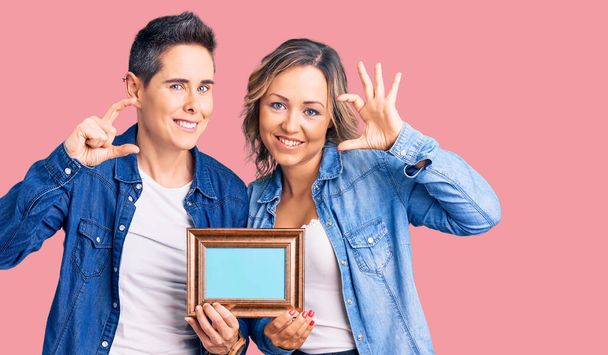 Couple de femmes tenant le cadre vide sourire et geste confiant avec la main faisant signe de petite taille avec les doigts à la recherche et la caméra. concept de mesure.  - Photo, image