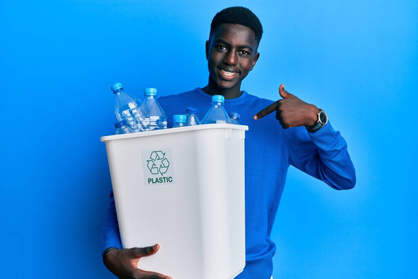 Mladý africký Američan držící recyklační odpadkový koš s plastovými lahvemi ukazující prstem na jednoho samolibě se usmívajícího šťastného a hrdého  - Fotografie, Obrázek