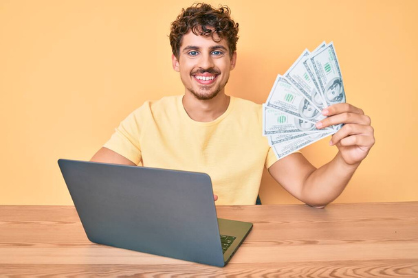 młody biały człowiek z kręconymi włosami działa za pomocą laptopa komputerowego trzymając usa dolarów banknotów patrząc pozytywnie i szczęśliwy stojąc i uśmiechając się z pewnym uśmiechem pokazując zęby  - Zdjęcie, obraz
