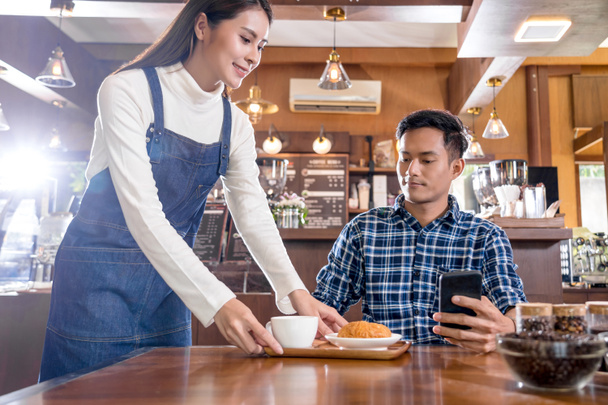 Asian Barista of Small business owner serving a cup of coffee and Croissants bakery to young customer at the table in coffee shop,Small business owner and startup in coffee shop and restaurant concept - Photo, Image