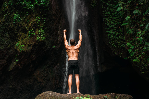 Back view of a man standing at a waterfall with his hands raised. The man at the waterfall raises his hands up. Man at the waterfall. Travel to Bali. Man on the background of nature. Copy space - 写真・画像