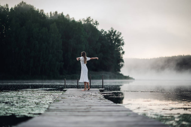 Вид ззаду на молоду жінку в білому платті, що стоїть на самоті на пішохідному мосту і дивиться в озеро. Туманний холодний ранок з туманом над водою
. - Фото, зображення