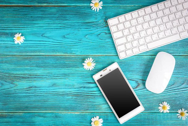 Table de travail d'été avec clavier, souris d'ordinateur, téléphone portable et fleurs de camomille sur table en bois bleu. - Photo, image