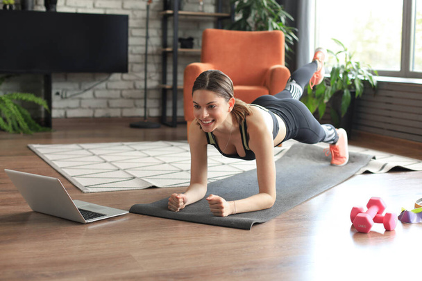 Beautiful fitness woman doing a plank exercise watching online tutorials on laptop, training in living room. Healthy lifestyle. Girl goes in for sports at home. - Фото, изображение