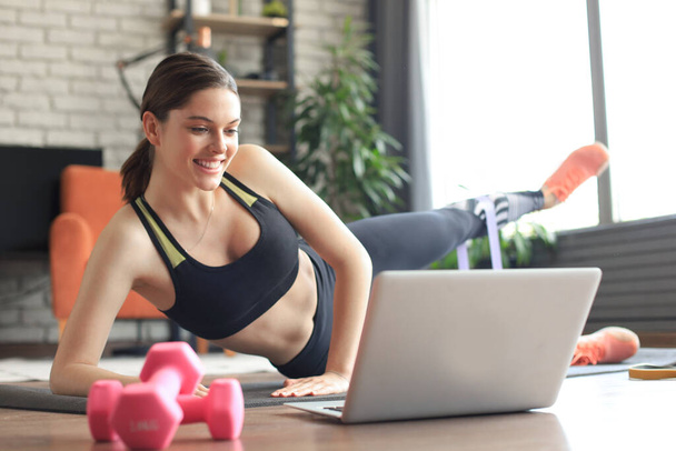 Fitness gyönyörű karcsú nő csinál oldalsó deszka egy rugalmas zenekar és néz online tutorials laptopon, edzés a nappaliban. Maradj otthon!. - Fotó, kép