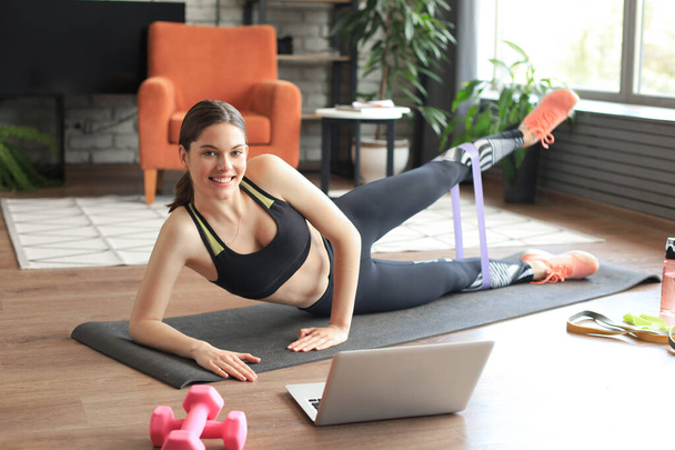 Fitness beautiful slim woman doing side plank with an elastic band and watching online tutorials on laptop, training in living room. Stay at home activities. - Foto, imagen