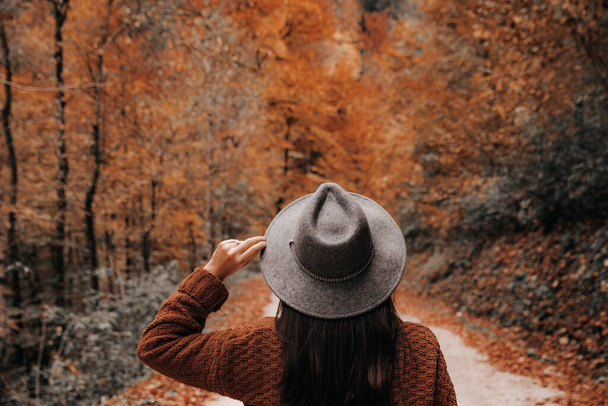 A shallow focus of a female wearing an orange sweater and a fedora hat and posing in a forest in autumn - Фото, зображення