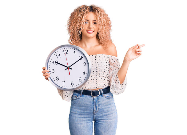 Young blonde woman with curly hair holding big clock smiling happy pointing with hand and finger to the side  - Фото, изображение