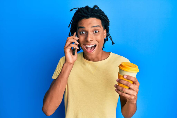 Jeune homme afro-américain utilisant un smartphone et boire une tasse de café célébrant fou et étonné pour le succès avec les yeux ouverts hurlant excité.  - Photo, image