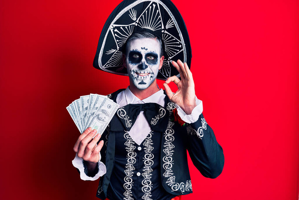 Young man wearing mexican day of the dead costume holding dollars doing ok sign with fingers, smiling friendly gesturing excellent symbol  - Φωτογραφία, εικόνα
