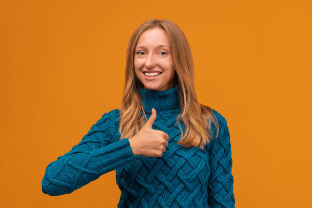 Mujer joven positiva en suéter azul de punto sonriendo y mostrando un gesto de pulgar hacia arriba. Buen trabajo, felicidad, lenguaje corporal, concepto de expresión facial. Estudio plano, fondo amarillo, aislado - Foto, Imagen