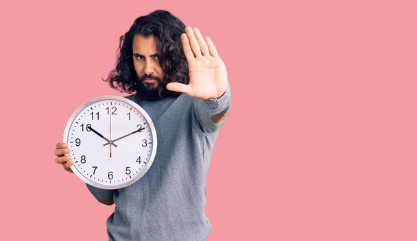 młody arabski człowiek trzyma duży zegar z otwartą ręką robi znak stopu z poważnym i pewnym siebie wyrazem twarzy, gest obrony  - Zdjęcie, obraz
