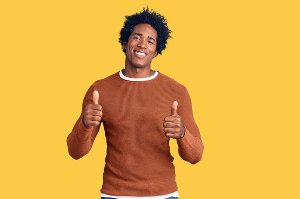 Schöner afrikanisch-amerikanischer Mann mit Afro-Haaren, lässig gekleidet und mit positiver Geste mit der Hand, Daumen hoch lächelnd und glücklich. Fröhlicher Ausdruck und Siegergeste.  - Foto, Bild