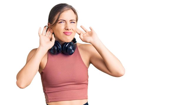 Krásná kavkazská mladá žena, oblečená do tělocvičny a používající sluchátka, která se snaží slyšet obě ruce na uchu, zvědavá na klepy. sluchový problém, hluchý  - Fotografie, Obrázek