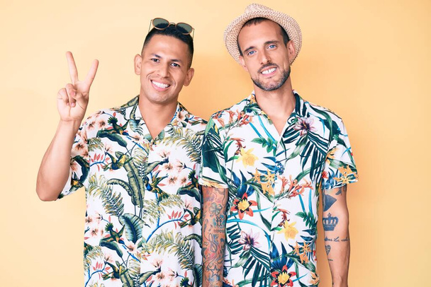 Молодая пара геев, одетых в летнюю шляпу и гавайскую рубашку, показывающих пальцами номер два, улыбающихся уверенно и счастливо.  - Фото, изображение