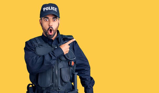 Giovane uomo ispanico indossa uniforme della polizia sorpreso indicando con il dito di lato, bocca aperta espressione stupita.  - Foto, immagini