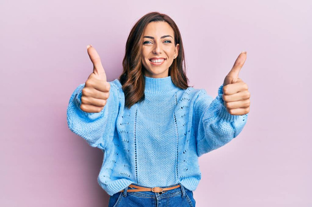 Mujer morena joven vistiendo suéter de invierno casual sobre fondo rosa aprobando hacer gesto positivo con la mano, pulgares arriba sonriendo y feliz por el éxito. gesto ganador.  - Foto, imagen