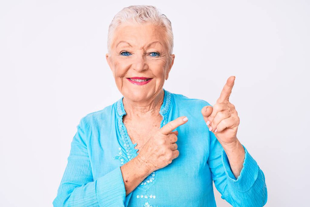 Vanhempi kaunis nainen, jolla on siniset silmät ja harmaat hiukset yllään kesävaatteet hymyillen ja katsoen kameraa osoittaen kahdella kädellä ja sormella sivulle.  - Valokuva, kuva