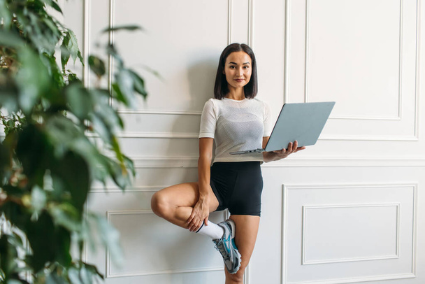 young beautiful girl with Asian appearance, fitness instructor, she conducts a web conference on fitness classes, she has a laptop and a phone in her hands - Foto, imagen