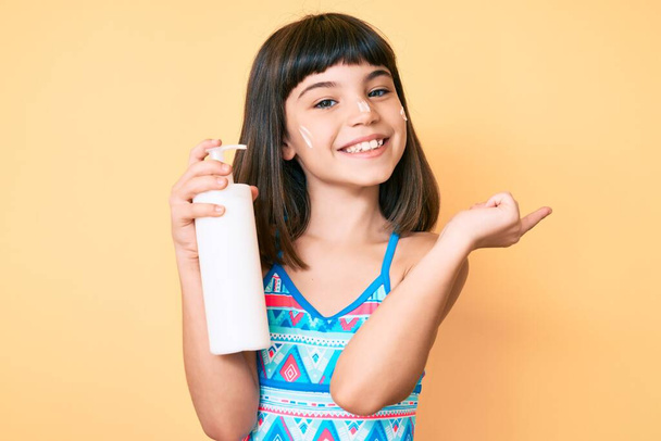 Νεαρό κοριτσάκι με bang φορώντας μαγιό και κρατώντας αντηλιακή λοσιόν χαμογελώντας χαρούμενος δείχνοντας με το χέρι και το δάχτυλο στο πλάι  - Φωτογραφία, εικόνα
