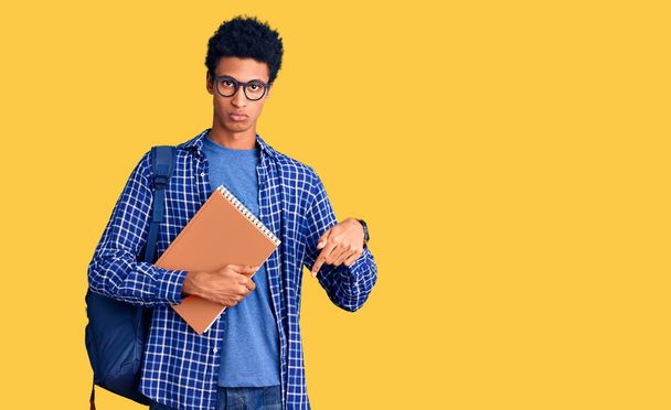Joven hombre afroamericano con mochila de estudiante sosteniendo libro apuntando hacia abajo mirando triste y molesto, indicando dirección con los dedos, infeliz y deprimido.  - Foto, imagen