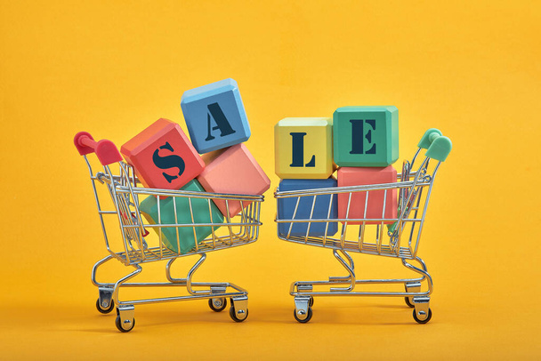 A miniature shopping cart and multi-colored cubes on yellow background. Sales word written on cubes. - Photo, Image