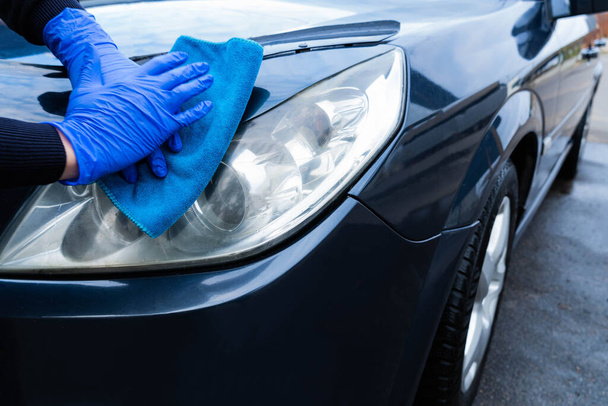 Een hand in een blauwe rubberen handschoen met een blauwe microvezel doek wrijft en polijst de koplamp van een auto. Was de auto zelf maar. Close-up - Foto, afbeelding